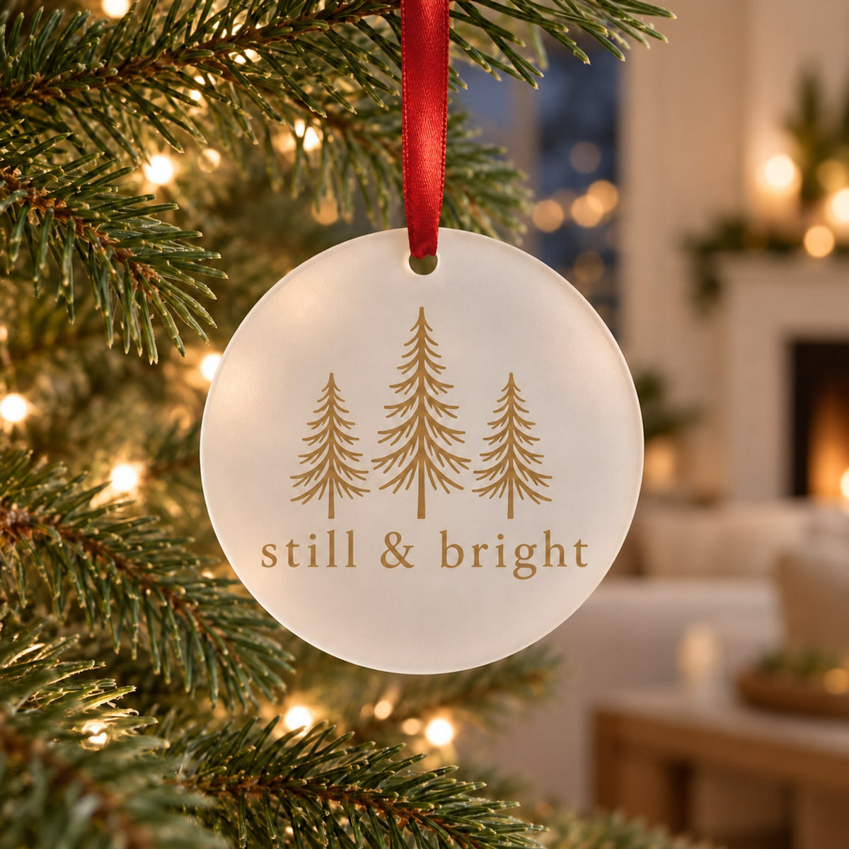 Frosted acrylic ornament with gold evergreen trees and still & bright hanging on a lit Christmas tree