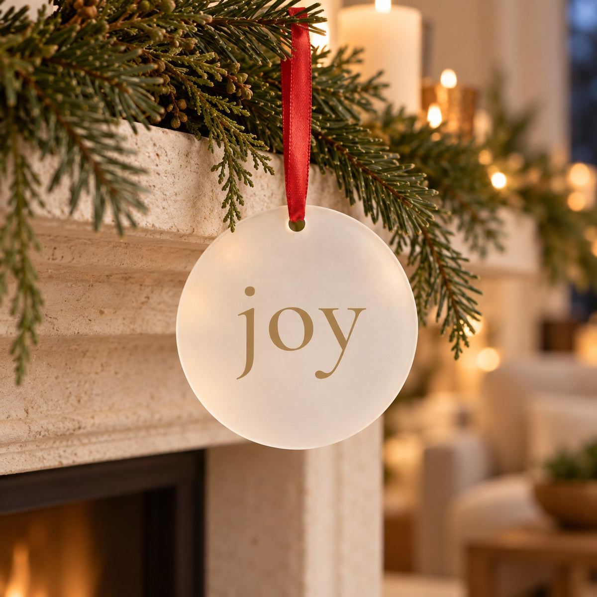 Frosted acrylic ornament with joy in champagne gold hanging on red ribbon from garland above a fireplace