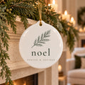 White ceramic ornament with a pine branch and noel hanging from garland on a candlelit mantel