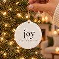 Hand hanging a white ceramic ornament printed with joy on a lit Christmas tree