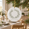 White ceramic ornament with a sage olive wreath hanging from a tree above a candlelit holiday table
