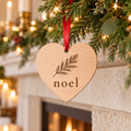 Wooden heart ornament with a pine branch and noel hanging from garland on a mantel with candles