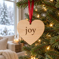 Wooden heart ornament printed with joy hanging on a red ribbon from a lit Christmas tree by a snowy window