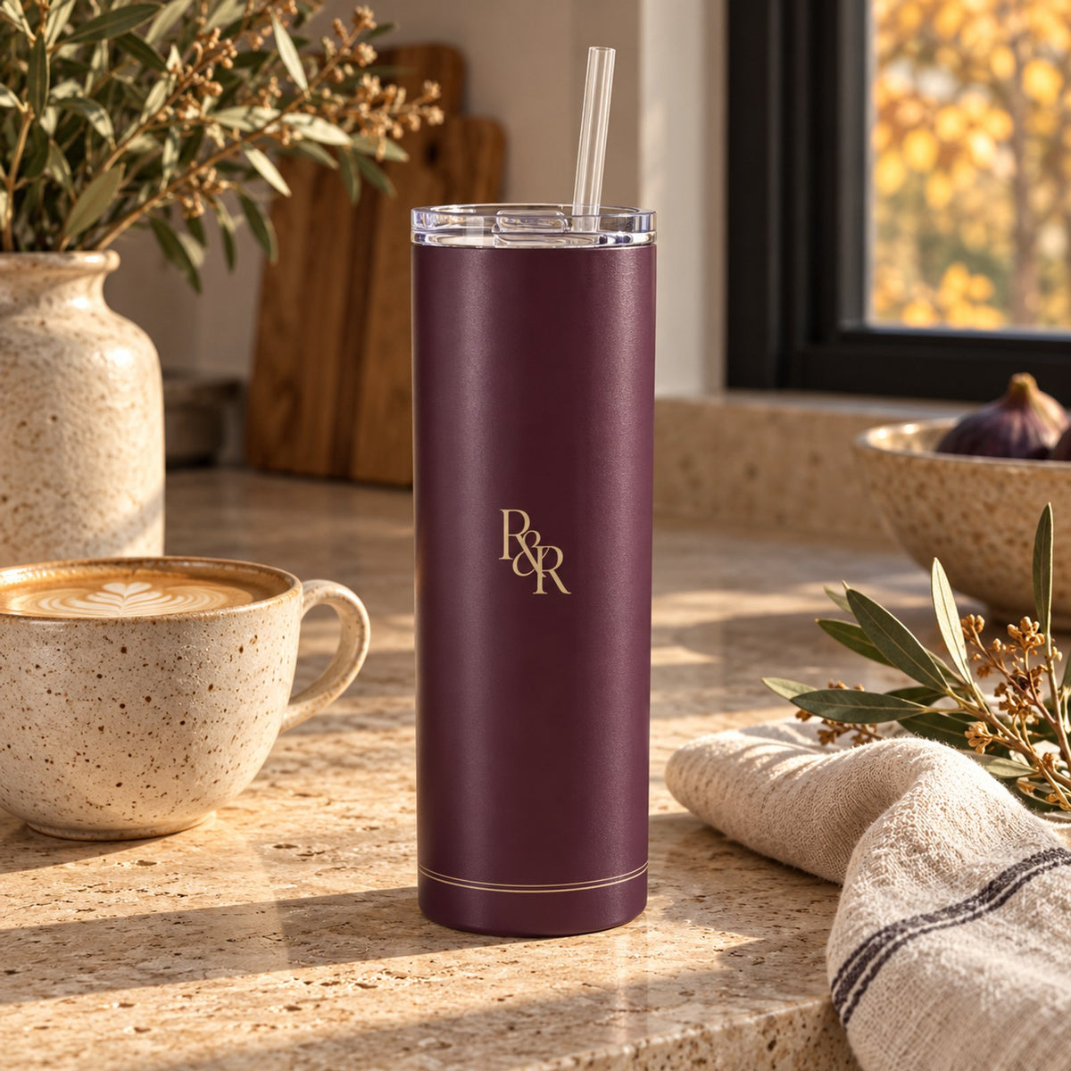 Deep fig tumbler with a champagne R&R monogram on a stone counter beside a latte and olive branches
