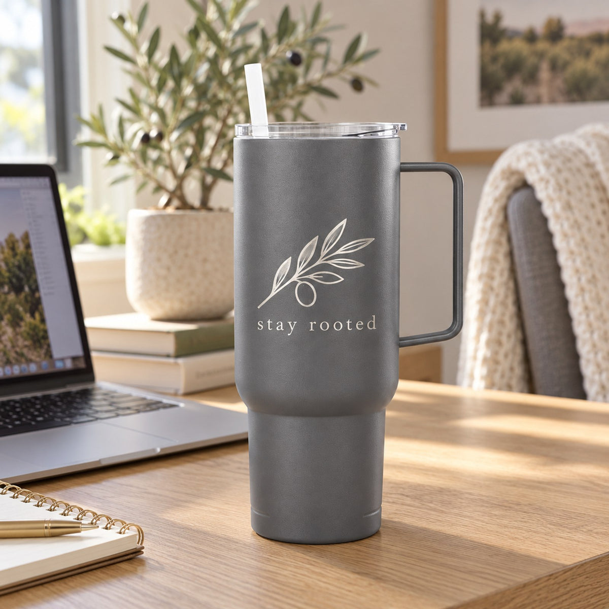 Gray 40 oz tumbler with handle engraved with an olive branch and stay rooted on a desk by a laptop