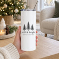 Hand holding a white skinny tumbler with evergreen trees and still & bright in a cozy holiday living room