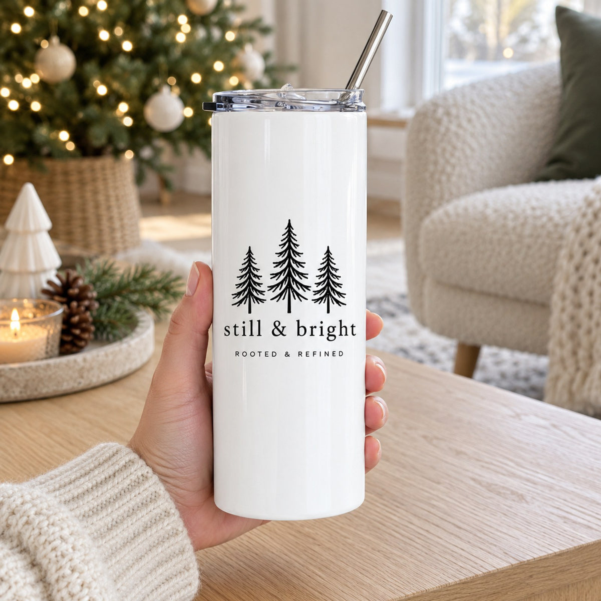 Hand holding a white skinny tumbler with evergreen trees and still & bright in a cozy holiday living room