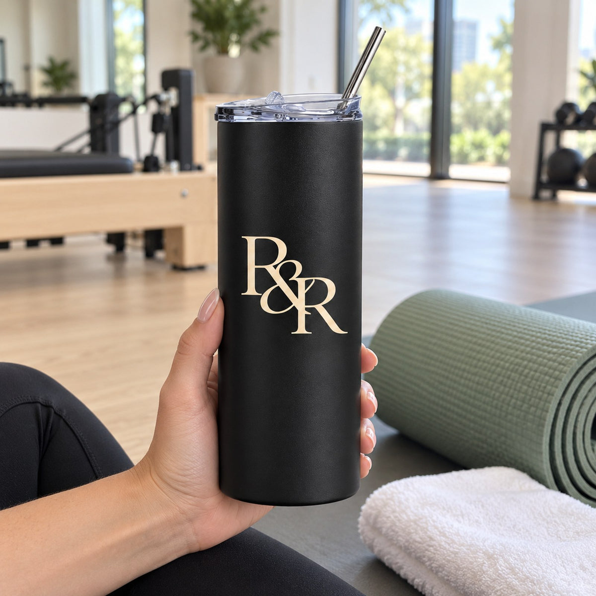 Hand holding a matte black skinny tumbler with a gold R&R monogram in a bright studio with a yoga mat
