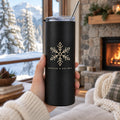 Hand holding a matte black skinny tumbler with a gold snowflake in a cozy living room by a fireplace