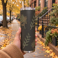 Hand holding a charcoal tumbler with sage and gold leaves on a tree-lined street covered in autumn leaves