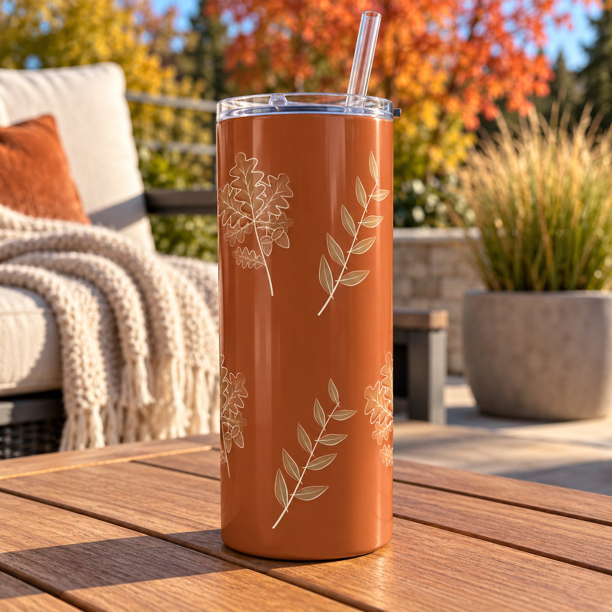 Rust tumbler with cream oak leaves and olive sprigs on an outdoor table with fall foliage and a throw