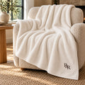 Ivory plush throw with a small espresso R&R monogram embroidered at the corner, draped over a cream boucle armchair