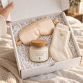 Open gift box with a champagne satin sleep mask, ivory lounge socks and a cork-lid candle with the cream Rooted & Refined label