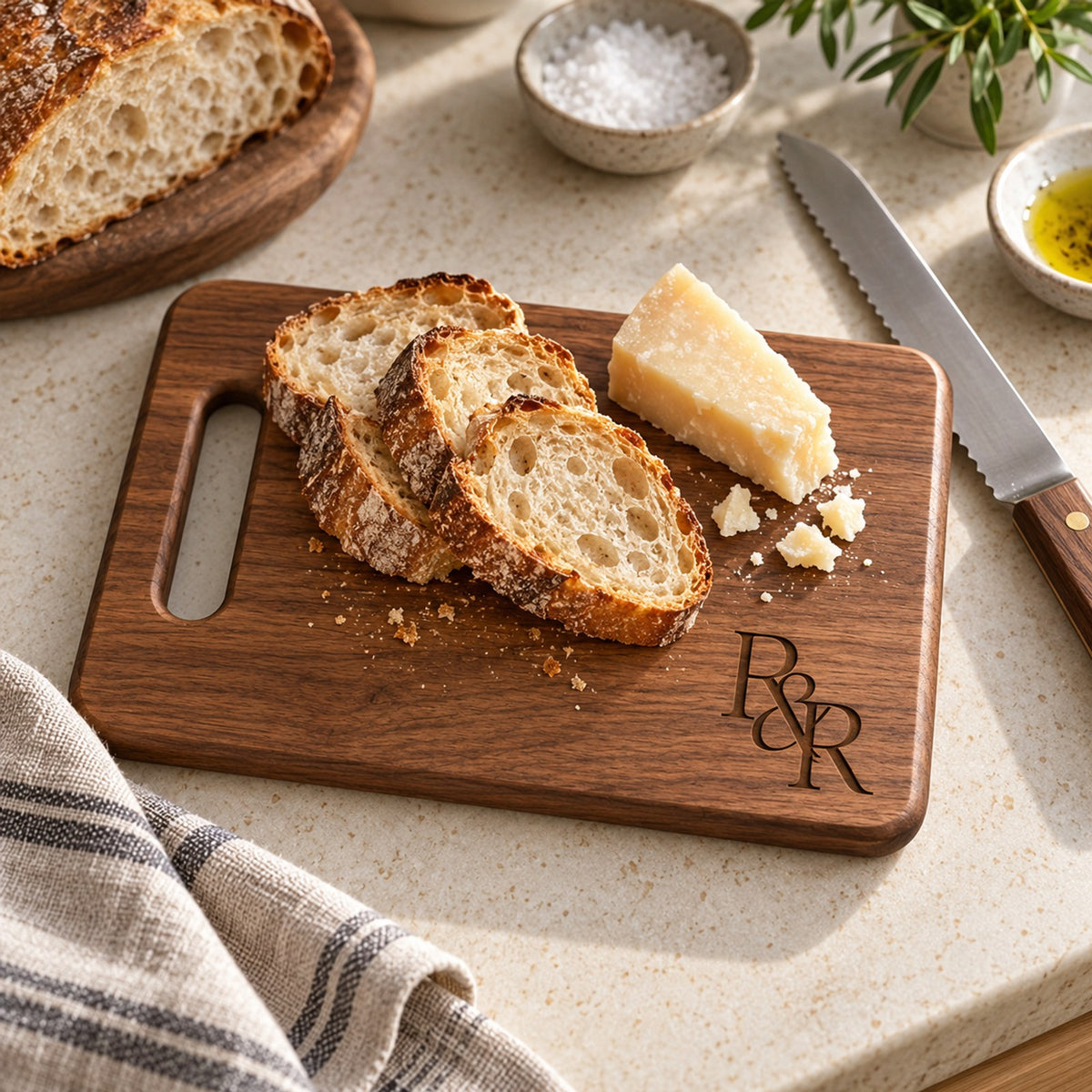 Walnut cutting board engraved with an R&R monogram, with sliced sourdough and parmesan on a stone counter