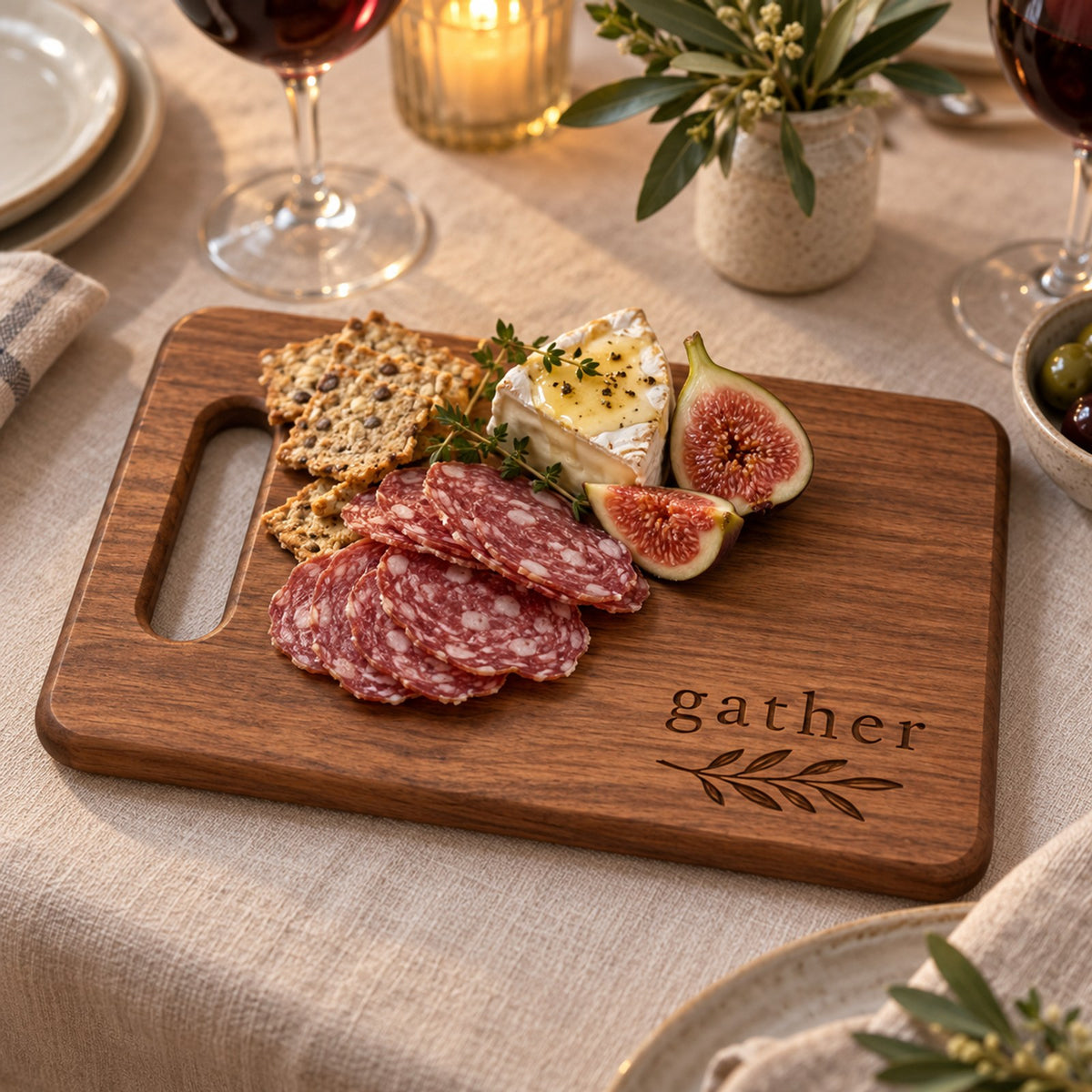 Walnut cutting board engraved with gather and an olive sprig, with salami, figs and brie on a candlelit table