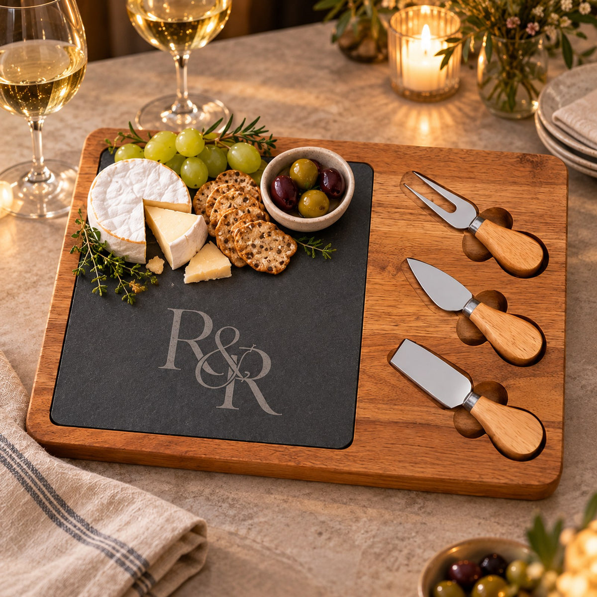 Acacia and slate charcuterie board engraved with an R&R monogram, with brie, grapes, olives and three cheese knives