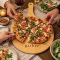 Bamboo pizza board engraved with gather, with friends sharing a prosciutto and arugula pizza