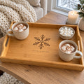Bamboo serving tray engraved with a snowflake, with mugs of cocoa and marshmallows by a snowy window