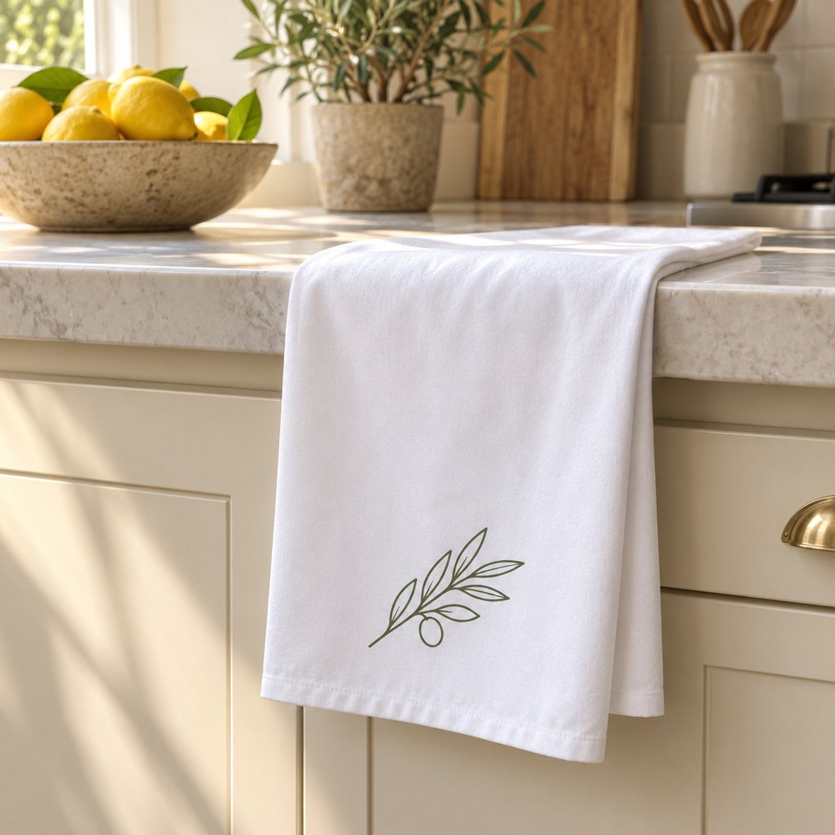 White tea towel with a sage olive branch draped over a kitchen counter beside a bowl of lemons