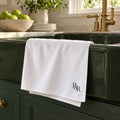 White tea towel with a black R&R monogram hanging on a deep green farmhouse sink