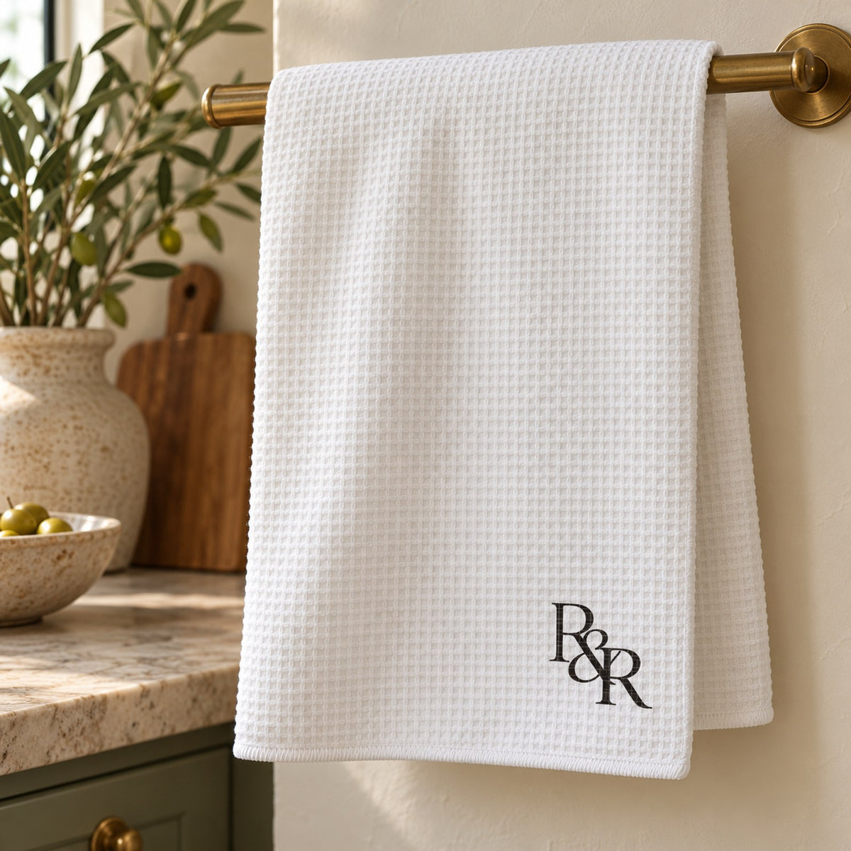 White waffle tea towel with a black R&R monogram hanging from a brass rail in a bright kitchen