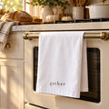 White tea towel printed with gather hanging on a brass oven rail in a fall kitchen with bread and pumpkins