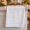 White tea towel with a gold gingerbread man draped on a counter beside trays of gingerbread cookies