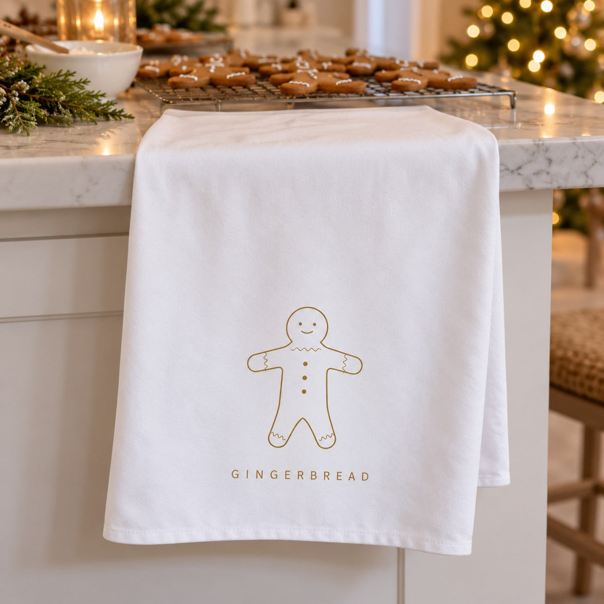 White tea towel with a gold gingerbread man draped on a counter beside trays of gingerbread cookies