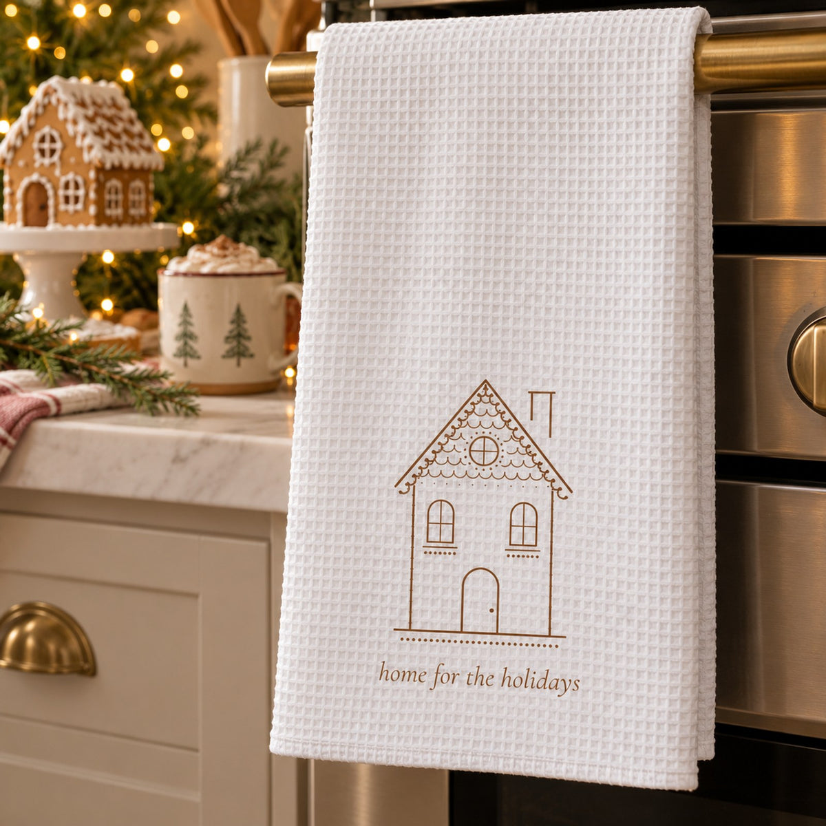 White waffle tea towel with a gingerbread house hanging on an oven beside a gingerbread house and cocoa