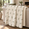 Ivory sherpa fleece blanket with an all-over olive branch pattern, draped over a cream sofa
