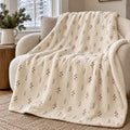 Linen-colour sherpa fleece blanket with a small winter berry sprig pattern, draped over a cream armchair