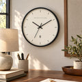 Signature ring wall clock with a black frame, cream face and fine hour marks, above a desk with a linen lamp