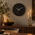Espresso-face wall clock with gold hour marks and a black frame, above a sideboard with brass candlesticks and pears