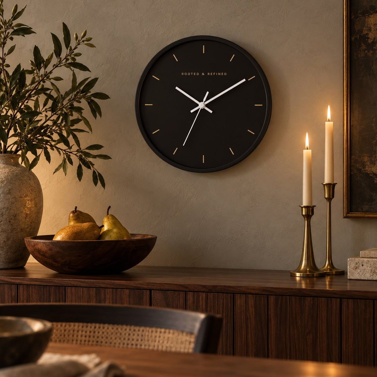 Espresso-face wall clock with gold hour marks and a black frame, above a sideboard with brass candlesticks and pears
