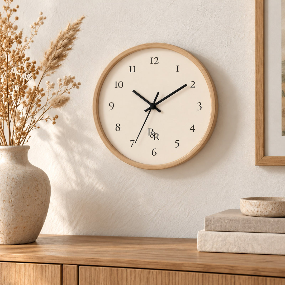 Round wall clock with a natural wood frame, cream face, numerals and a small R&R monogram, above a console with dried grasses