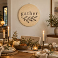 Round light wood sign printed with gather and an olive branch, hanging above a candlelit fall dining table