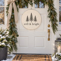 Round light wood sign with three evergreen trees and still & bright, hanging on a white front door with a garland