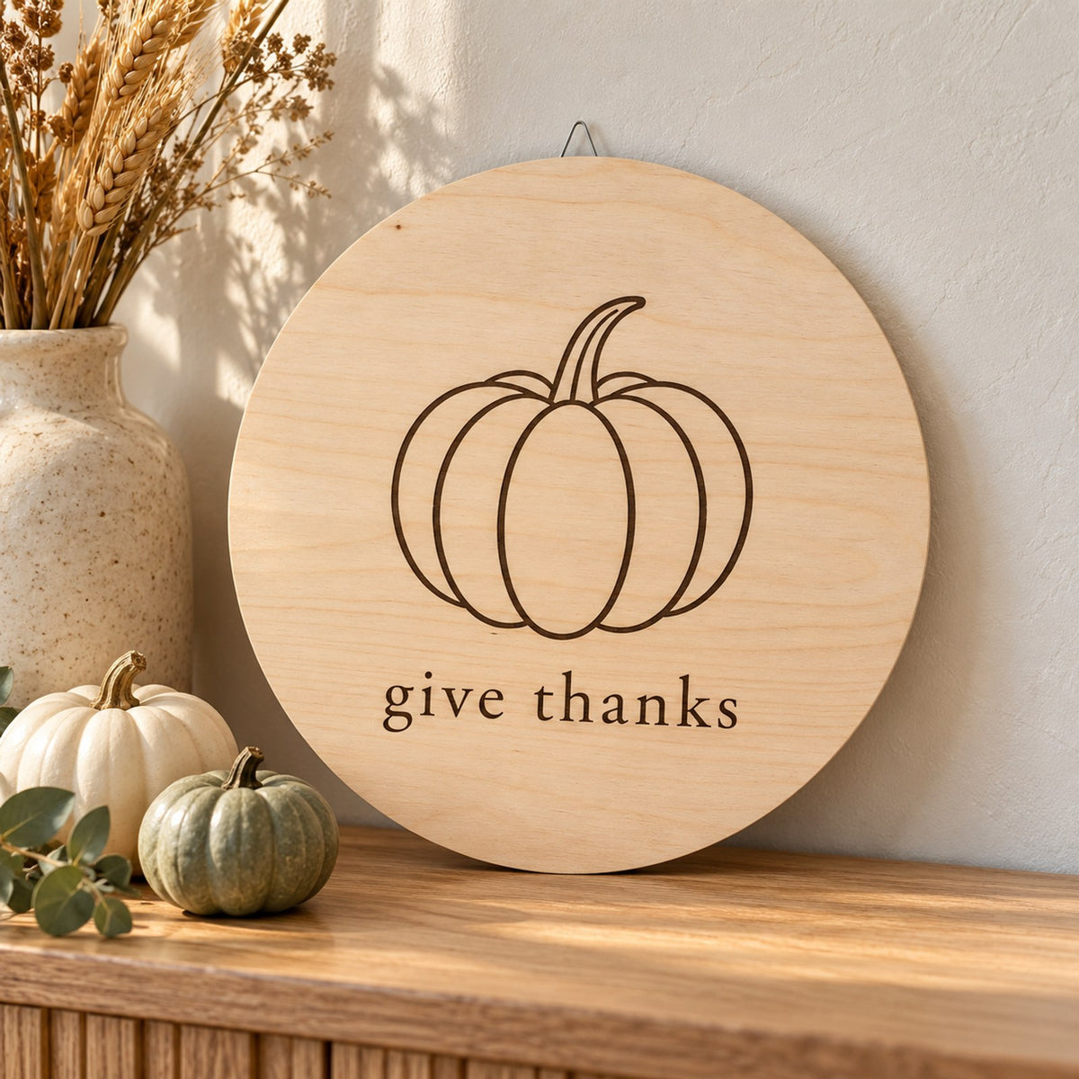 Round light wood sign with a line-drawn pumpkin and give thanks, leaning on a shelf with white and green pumpkins