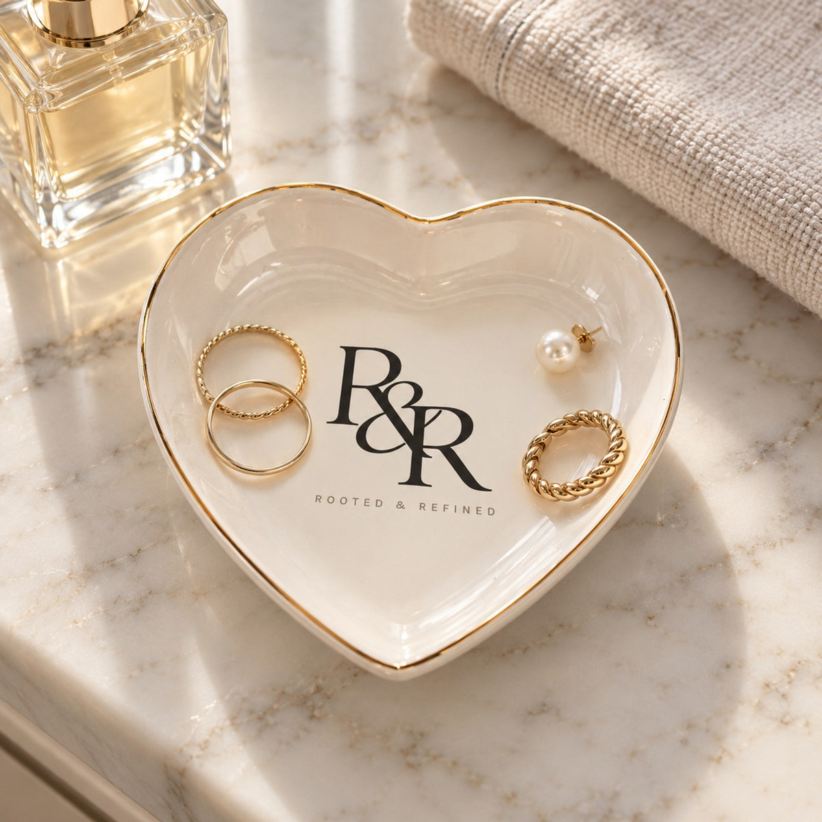 Gold-rimmed white heart trinket dish with the R&R monogram, holding gold rings and pearl earrings on marble