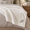 Ivory plush throw with a small embroidered olive branch at the corner, folded across a neutral linen bed