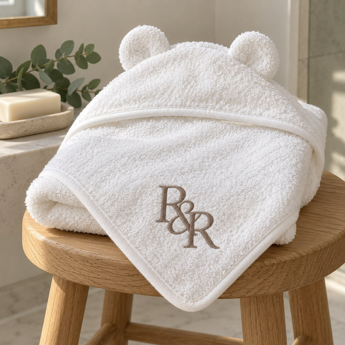 White bear-ear baby hooded towel with a taupe embroidered R&R monogram, folded on a wooden stool
