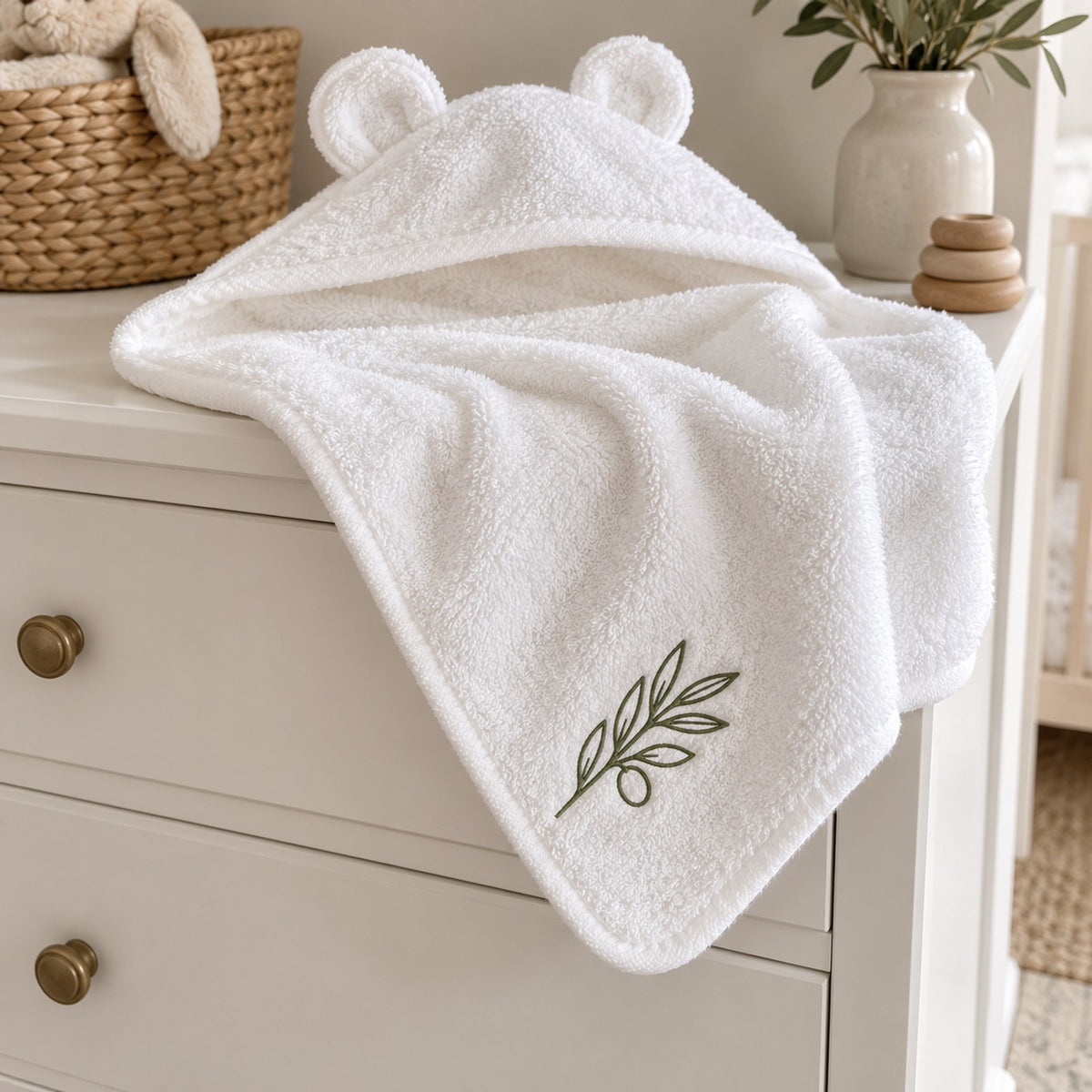 White bear-ear baby hooded towel with an embroidered olive branch, on a white nursery dresser