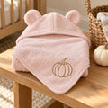 Pink bear-ear baby hooded towel with a camel embroidered pumpkin, on a wooden bench beside small white pumpkins