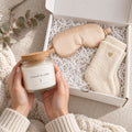 Hands holding a rooted in calm candle beside a gift box with a champagne satin sleep mask and ivory socks