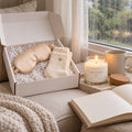 Gift box with a champagne satin sleep mask and ivory socks beside a lit a quiet moment candle and an open journal by a rainy window