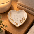 Gold-rimmed white heart trinket dish with the Rooted & Refined signature heart, holding gold hoops and a chain on a nightstand