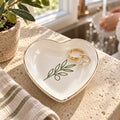 Gold-rimmed white heart trinket dish with an olive branch, holding gold rings beside a kitchen sink