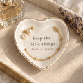 Gold-rimmed white heart trinket dish reading keep the little things, holding pearl earrings and a gold bracelet