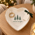 Gold-rimmed white heart trinket dish with three evergreen trees and still & bright, holding a gold ring with a gift tag