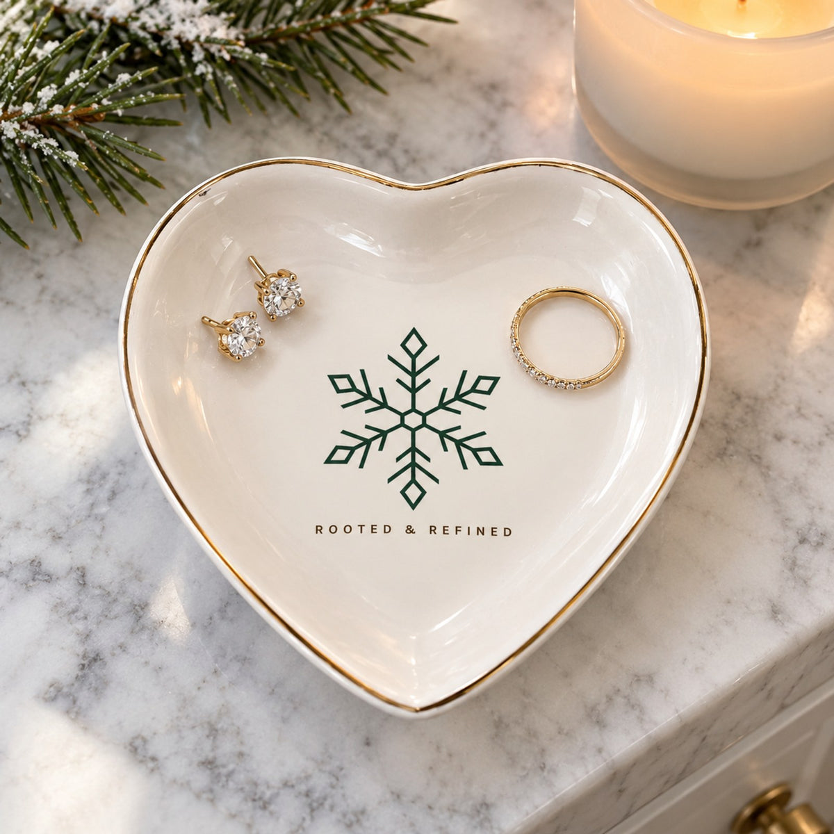 Gold-rimmed white heart trinket dish with a green snowflake, holding a ring and earrings on marble with pine